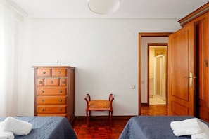 A room with two beds, a wooden dresser, a chair, and a door leading to another room. - 1A130 Coqueto apartamento cerca playa (Gijón)