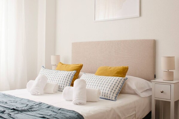 Una cama bien hecha con sábanas blancas, dos almohadas con un diseño azul y una almohada amarilla. Hay una mesita de noche con una lámpara y un cajón.