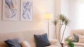 Een woonkamer met een beige bank, twee kussens, een vloerlamp en twee planten in potten.