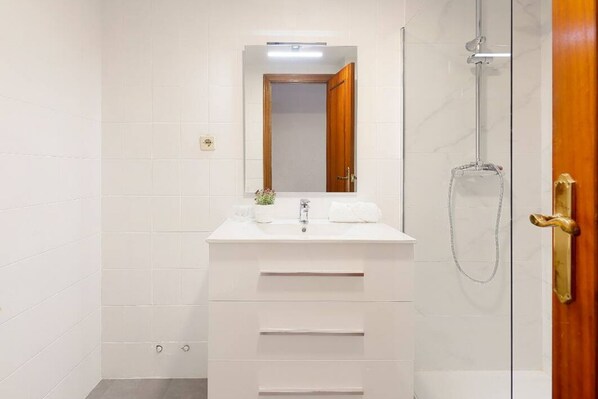 Un baño con tocador blanco, un espejo, ducha y puerta de madera.