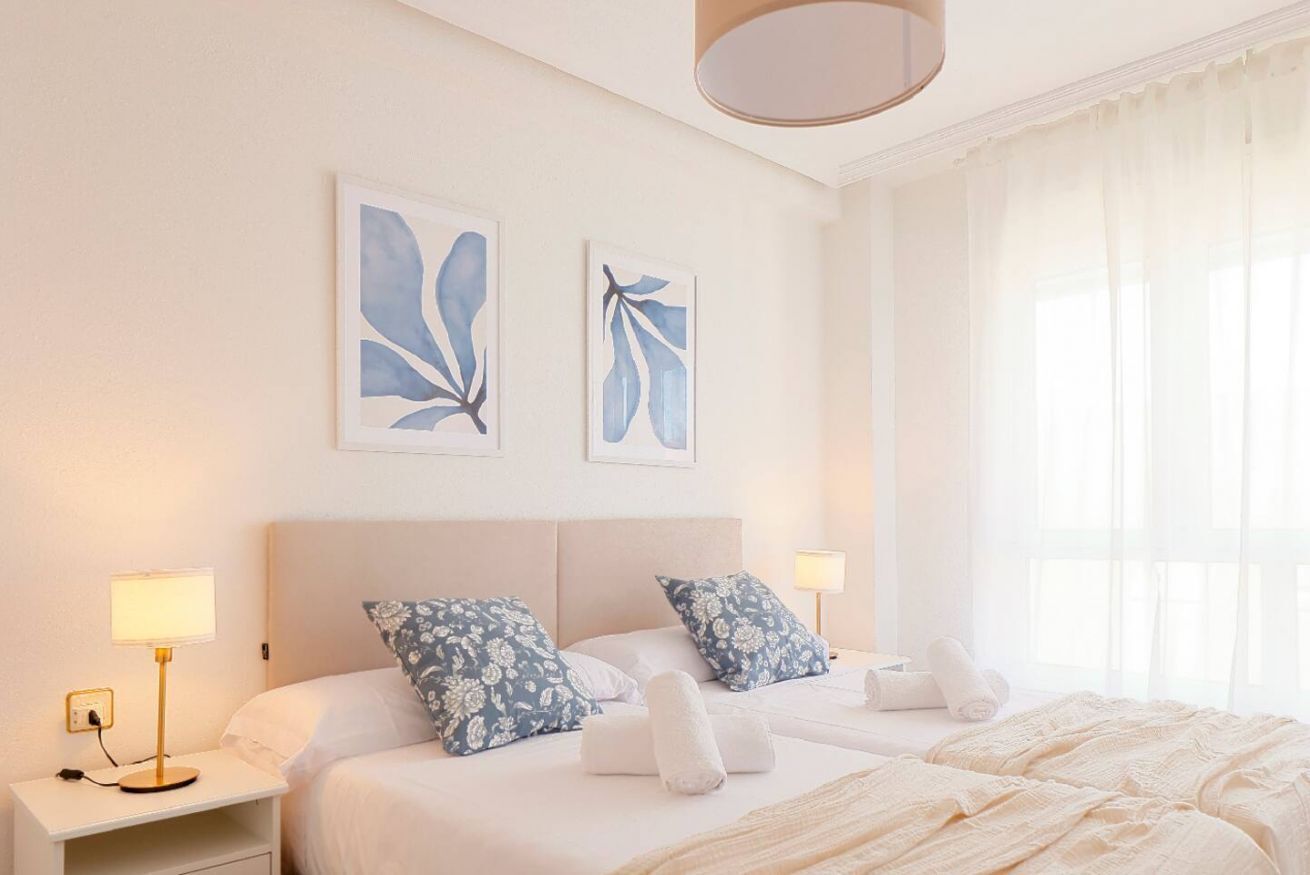 A hotel room with two beds, white bedding, floral-patterned pillows, artwork on the wall, and a bedside table with a lamp.