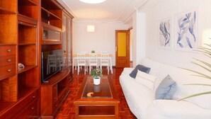 A living room with a sofa, coffee table, TV, wooden cabinets, and a dining area with chairs and a table. - 1A130 Coqueto apartamento cerca playa (Gijón)