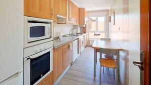 A kitchen with wooden cabinets, a microwave, an oven, and a dining area with a table and chair. - 1A130 Coqueto apartamento cerca playa (Gijón)