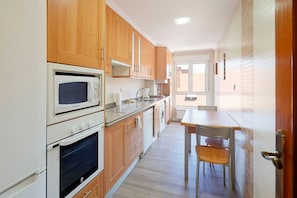 A kitchen with wooden cabinets, a microwave, an oven, and a dining area with a table and chair. - 1A130 Coqueto apartamento cerca playa (Gijón)