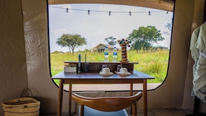 A tent with a table and chairs, a basket, and a view of a house and trees outside. - Malaika Mara River Luxury Camp (Serengeti)