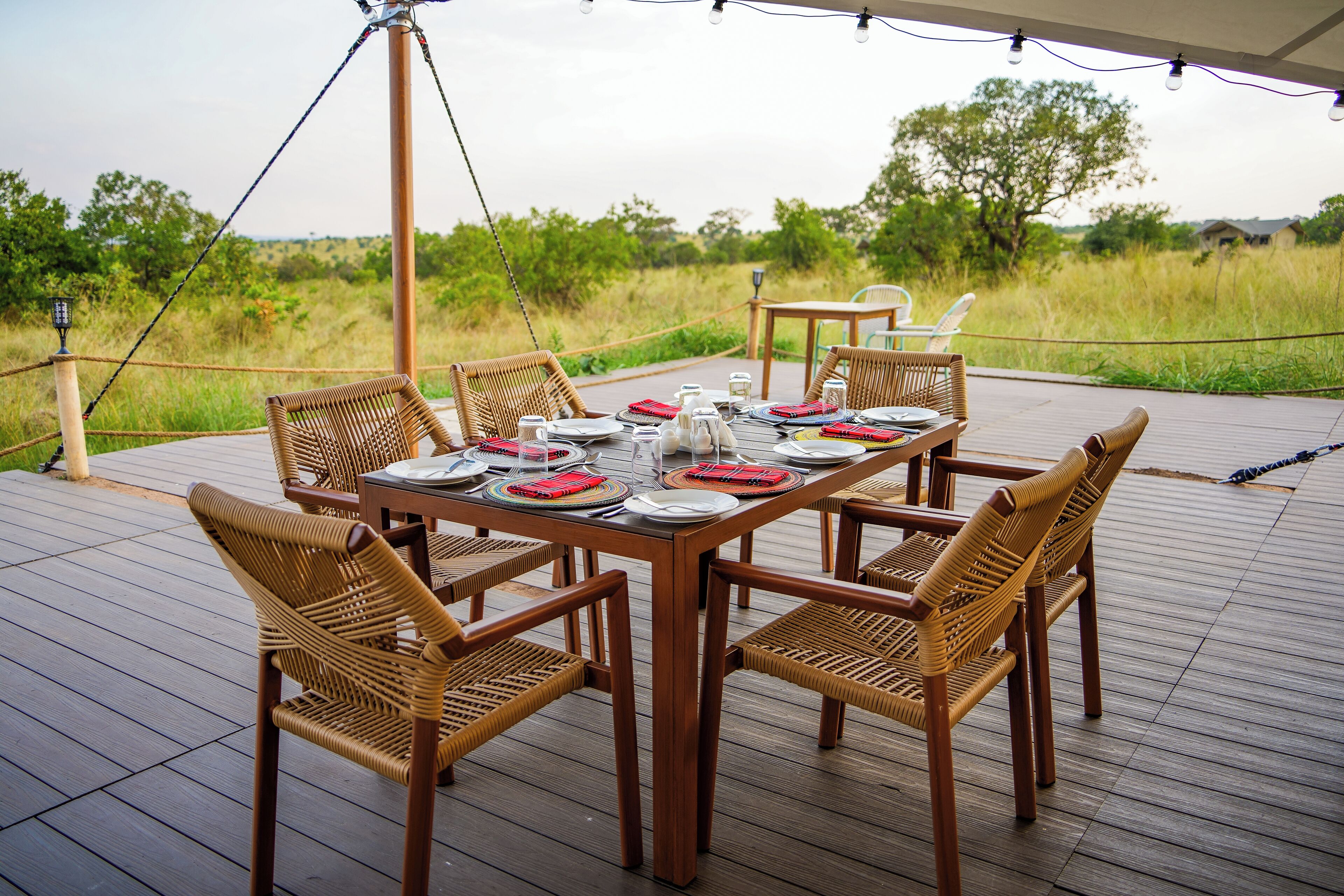 A wooden deck with a table set for six, surrounded by wicker chairs, overlooking a grassy field.
