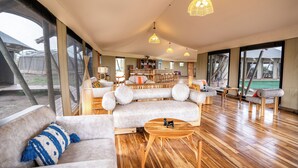 A spacious living area with a large sofa, a coffee table, and a dining table. There are multiple hanging lights and large windows providing natural light. - Malaika Mara River Luxury Camp (Serengeti)