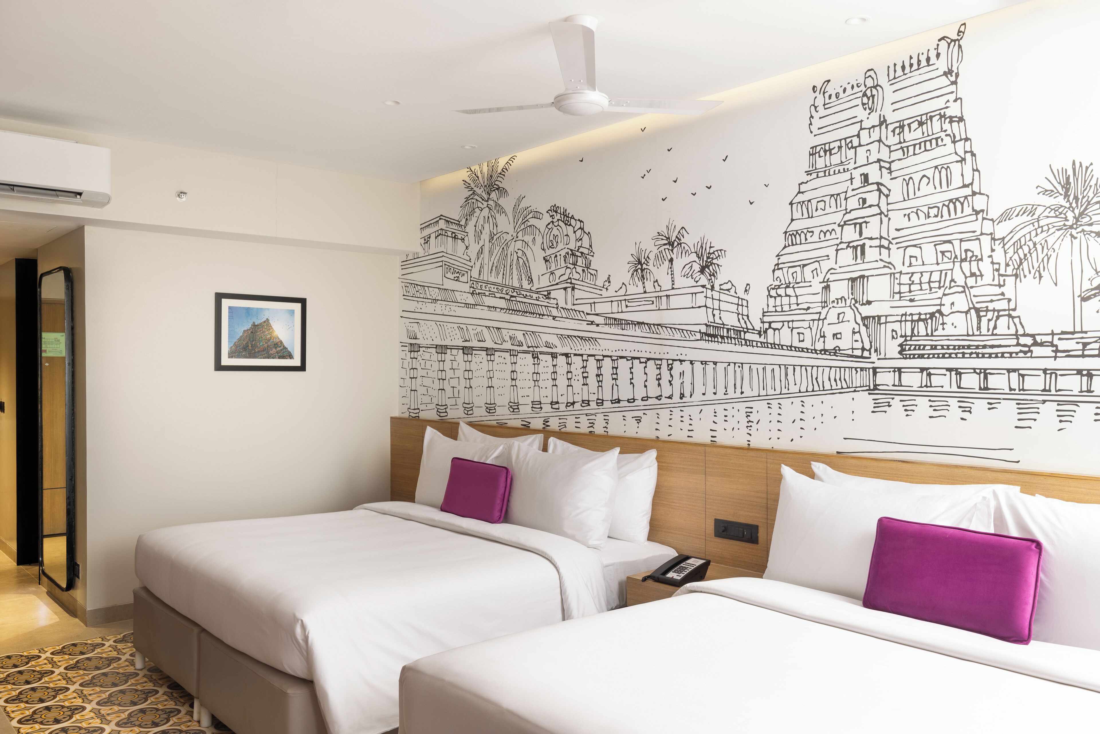 Une chambre d’hôtel avec deux lits, un ventilateur de plafond, une fresque murale et un tableau encadré.