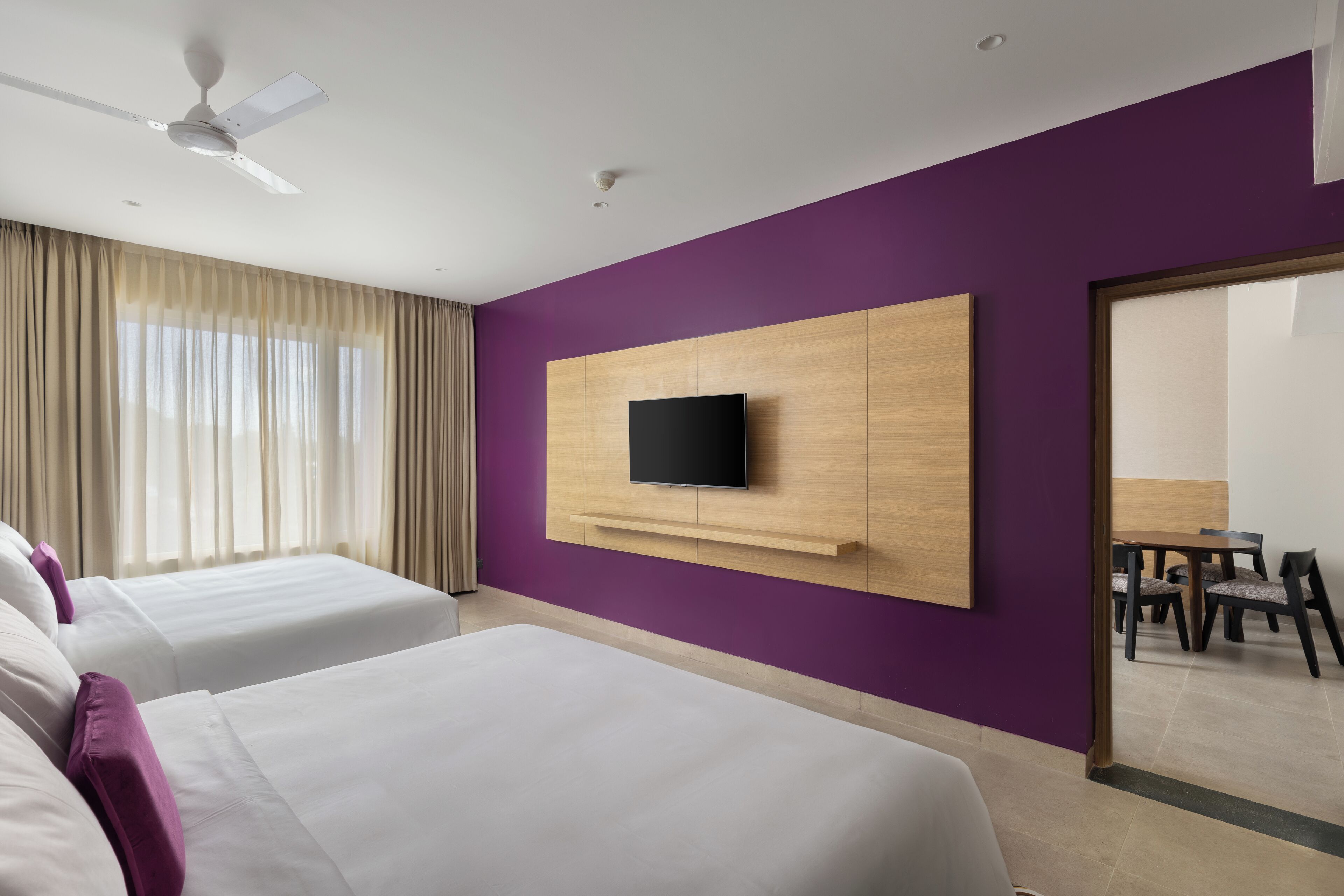 A hotel room with two beds, a TV mounted on a wooden panel, a ceiling fan, and a view of a dining area through a glass door.