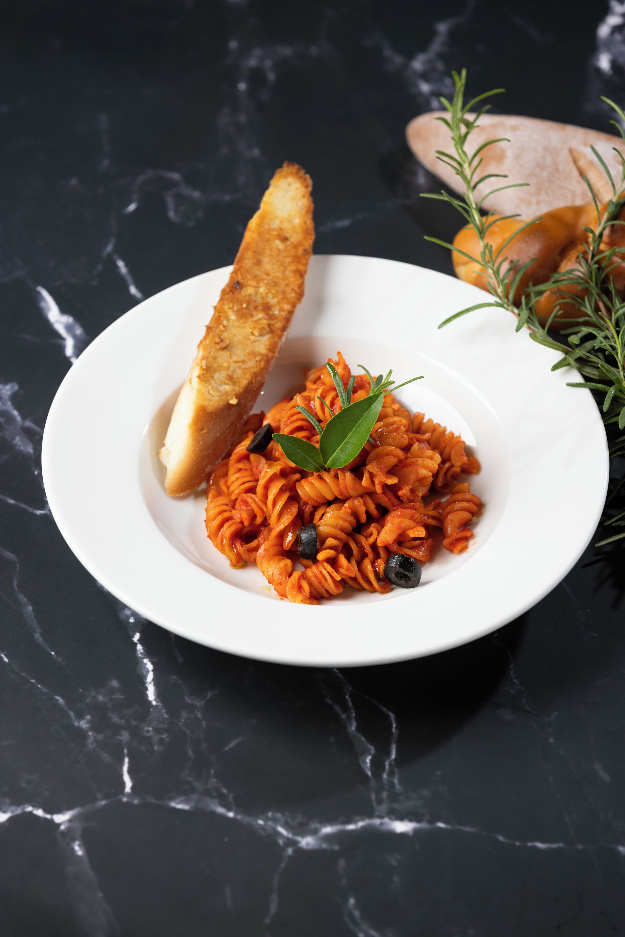 A plate of pasta garnished with a bay leaf and black olives, accompanied by a piece of bread on a dark marble surface.