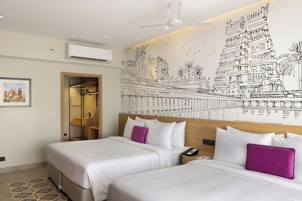 Habitación de hotel con dos camas, un ventilador de techo, un equipo de aire acondicionado, un mural en la pared y una puerta que da a un baño.