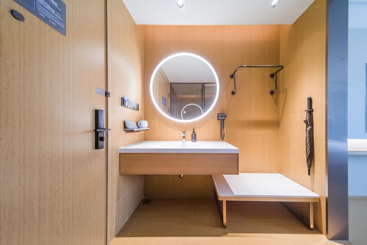A modern bathroom with a wooden vanity, a round mirror with lighting, a handheld showerhead, and a towel rack.