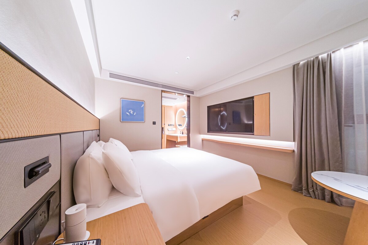 A modern hotel room with a large bed, a desk, a flat-screen TV, and a bathroom visible through an open door.
