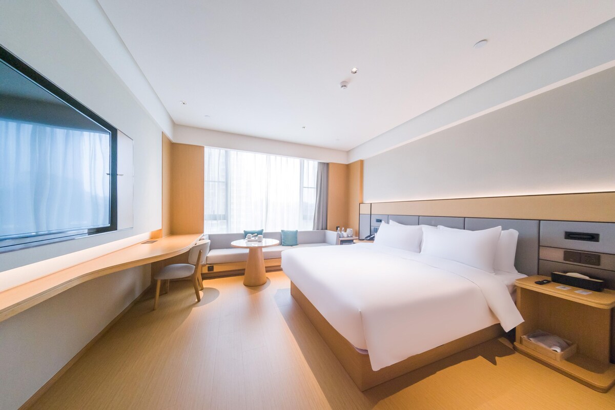 A modern hotel room with a large bed, a desk, a chair, a small round table, and a nightstand.