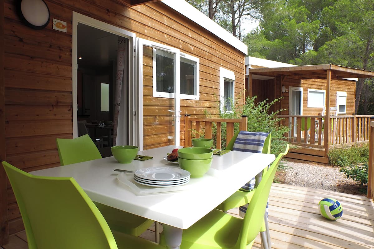Uma cabana de madeira com sala de jantar, com uma mesa branca e cadeiras verdes, um deck com uma bola de vôlei e um pequeno alpendre.