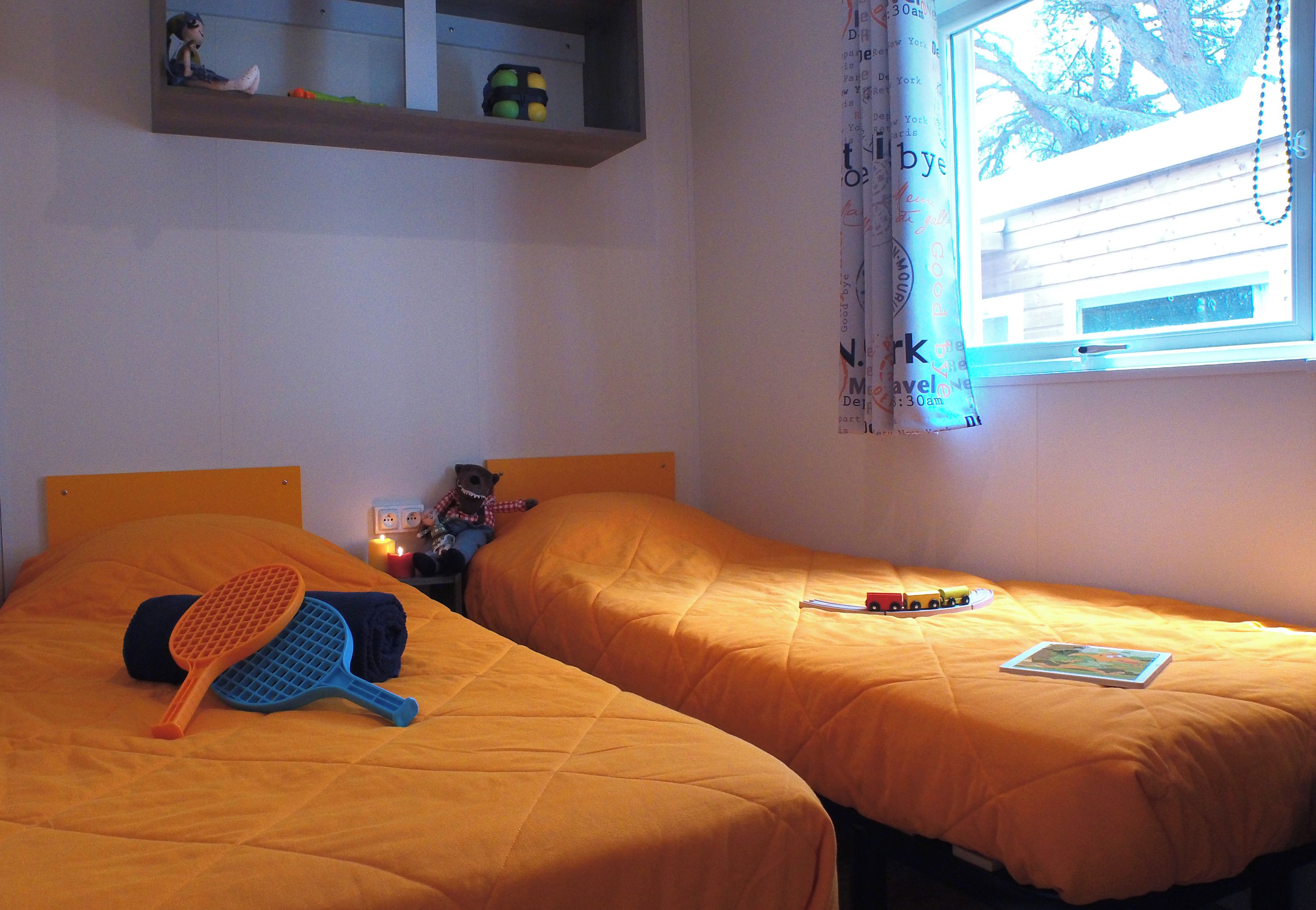 Ein Zimmer mit zwei Betten, einer Ablage mit Gegenständen, einem Fenster mit Vorhängen und einem kleinen Tisch mit einem Spielzeug.