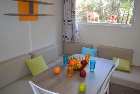 Una habitación con una mesa de comedor dispuesta con frutas y vasos, un sofá con cojines, y una ventana que ofrece vista a árboles y una estructura roja en el exterior.