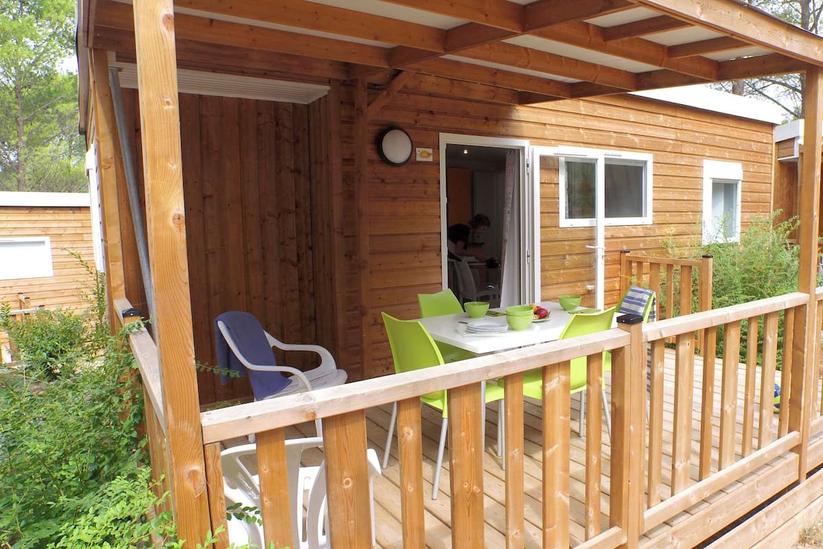 Una terraza de madera con una mesa, sillas, un ventanal y una puerta que da a un espacio interior.