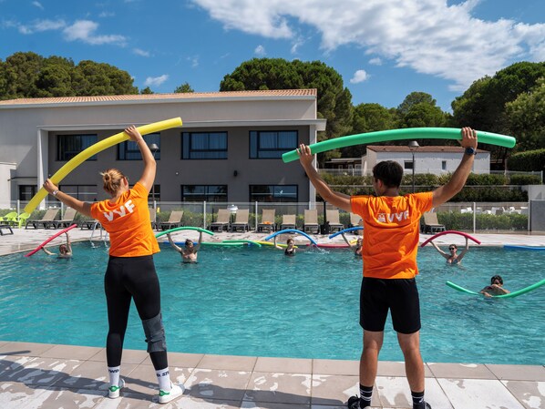 Zona de piscina con gente disfrutando de actividades acuáticas, un edificio con tejado de cerámica y cielo despejado.