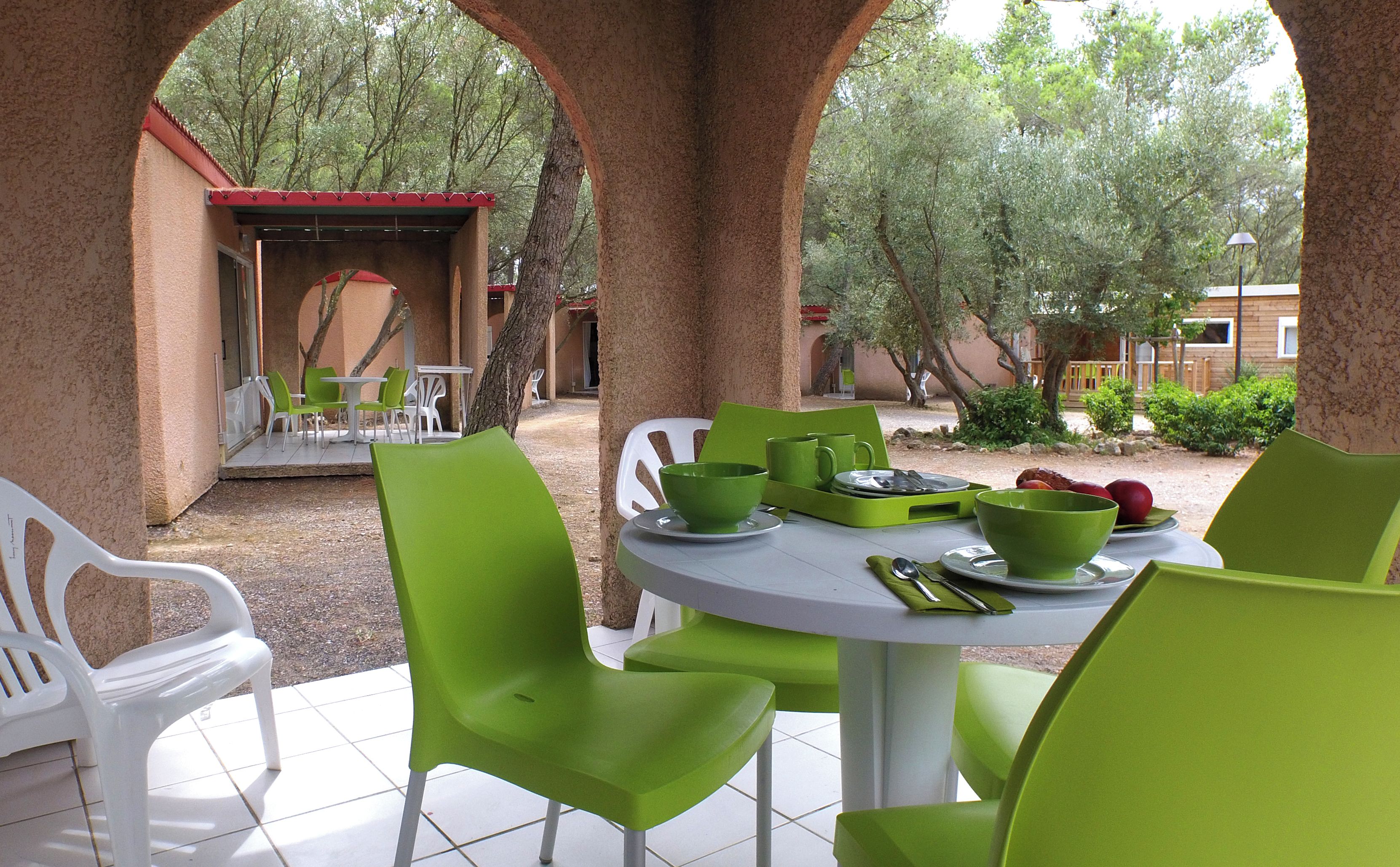 Un espace extérieur aménagé pour prendre les repas, avec des chaises vertes et des tables blanches, entouré d’arbres et d’un bâtiment doté d’un toit rouge.