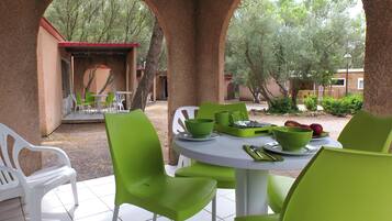 Un espace extérieur aménagé pour prendre les repas, avec des chaises vertes et des tables blanches, entouré d’arbres et d’un bâtiment doté d’un toit rouge.