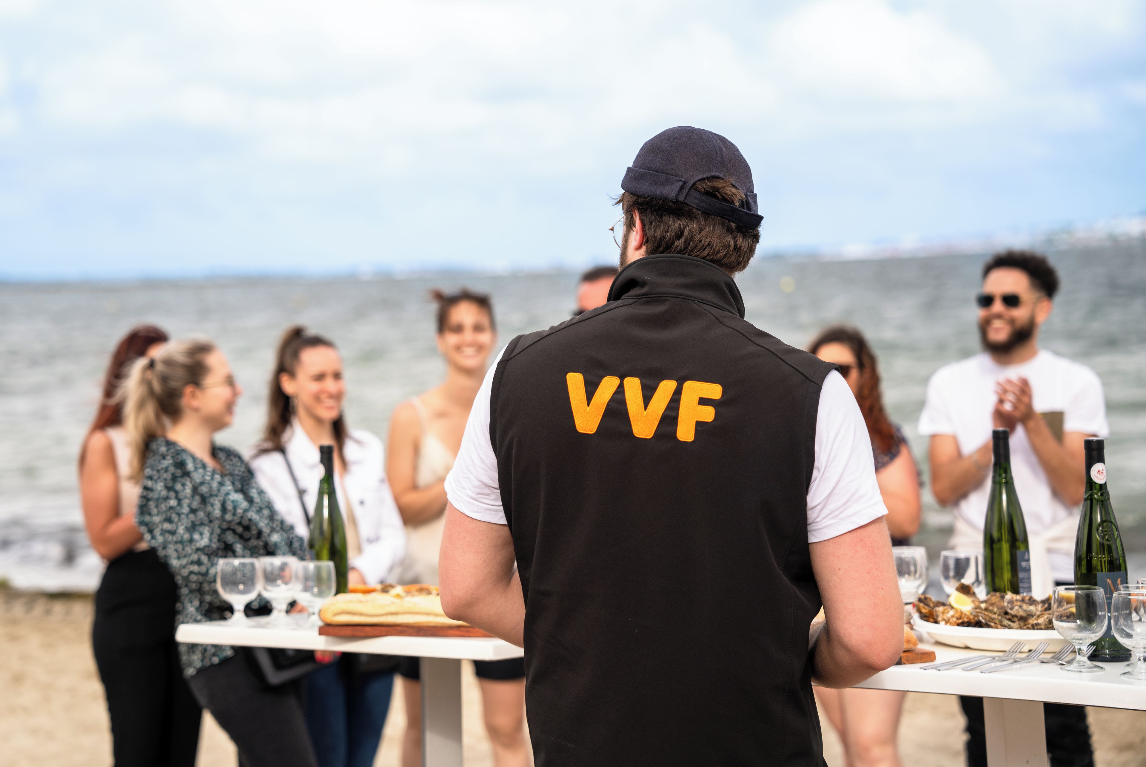 A person in a black vest with 'VVF' on the back standing at a table with a view of the sea in the background.