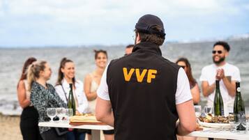A person in a black vest with 'VVF' on the back standing at a table with a view of the sea in the background.
