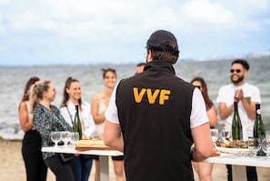 A person in a black vest with 'VVF' on the back standing at a table with a view of the sea in the background. - VVF Villages Montagnac (Montagnac)