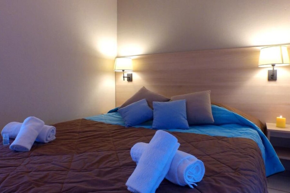 Una habitación de hotel con cama, almohadas, una mesita de noche con lámpara y toallas dobladas sobre la cama.