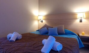 Una habitación de hotel con cama, almohadas, una mesita de noche con lámpara y toallas dobladas sobre la cama.