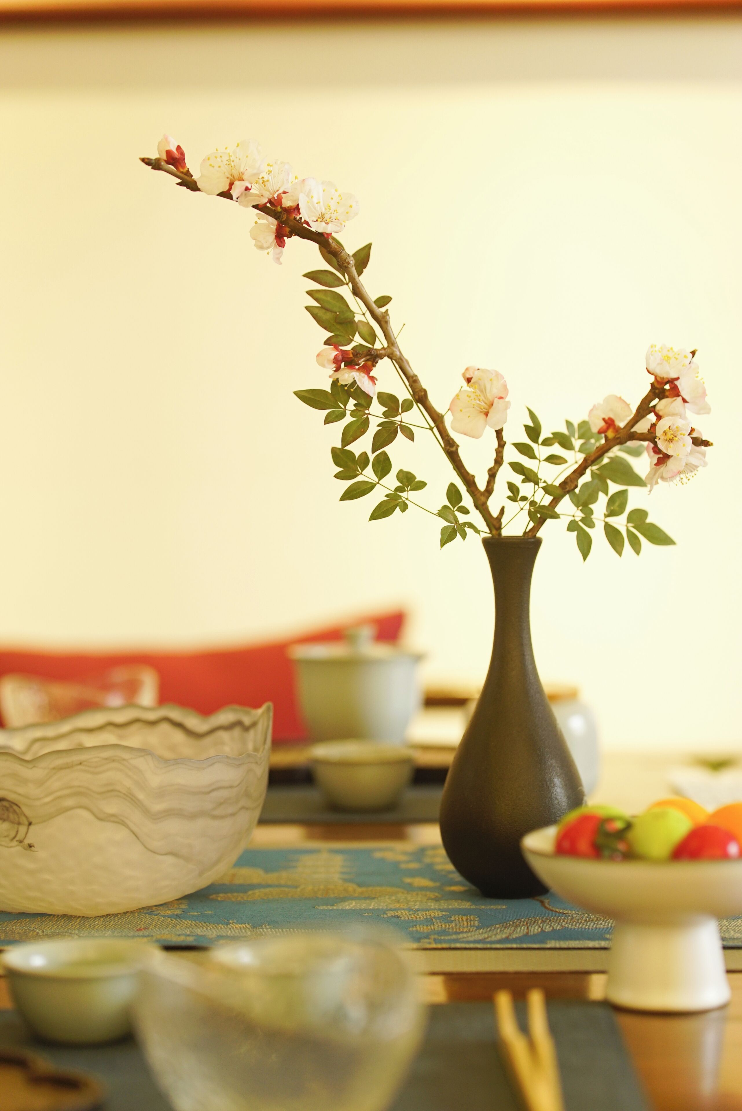 A dining table with a vase of flowers, a bowl, and a plate of fruit.