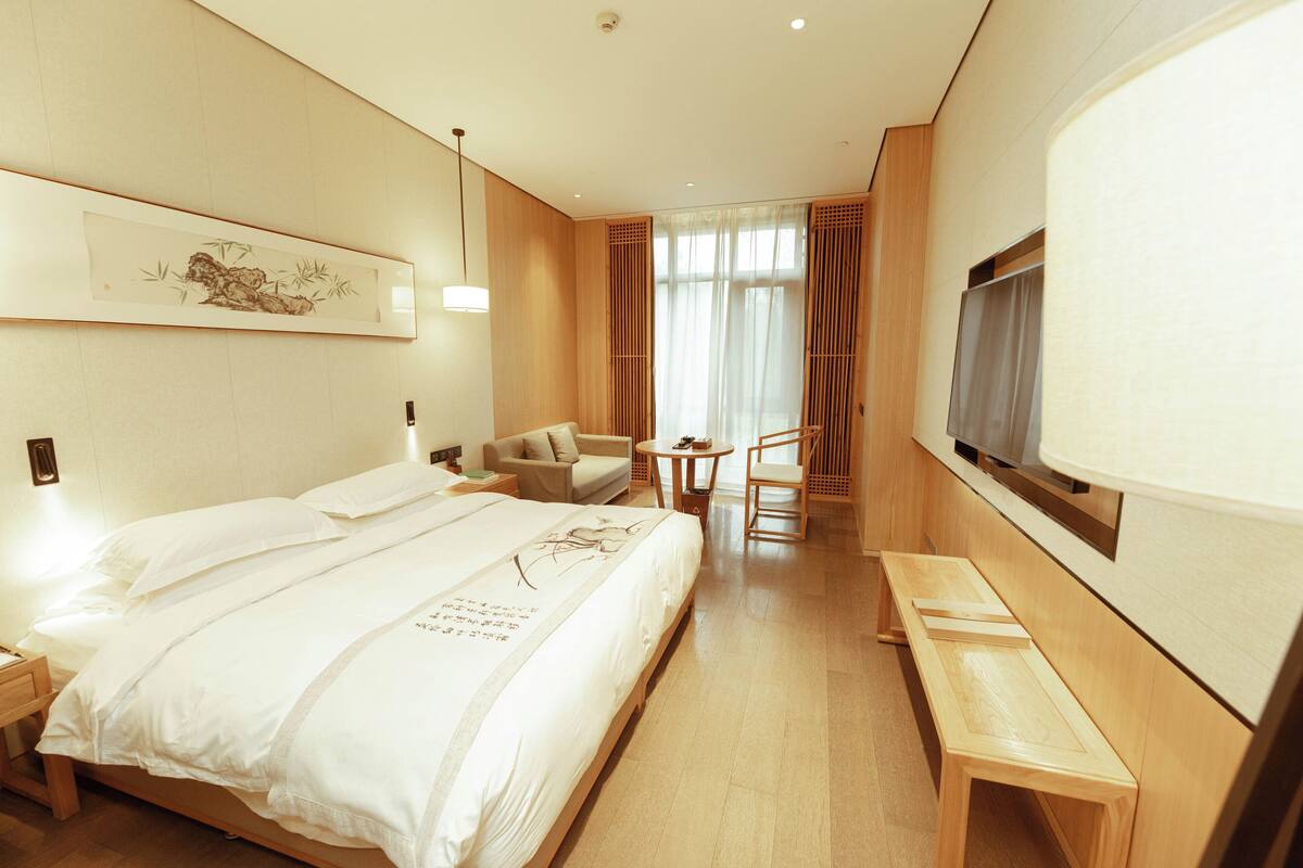 A hotel room with a large bed, a desk, a chair, a small table, and a TV mounted on the wall.