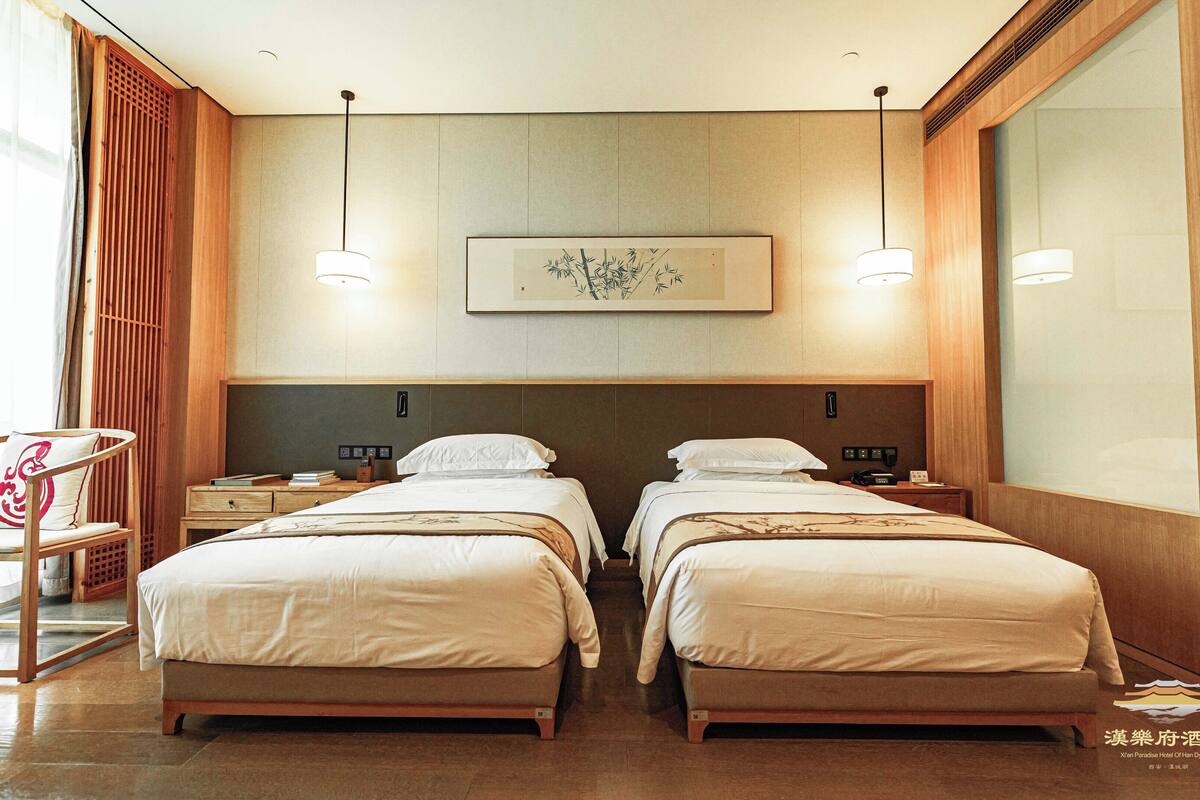 A hotel room with two beds, a wooden headboard, a small table with a chair, and a framed artwork on the wall.
