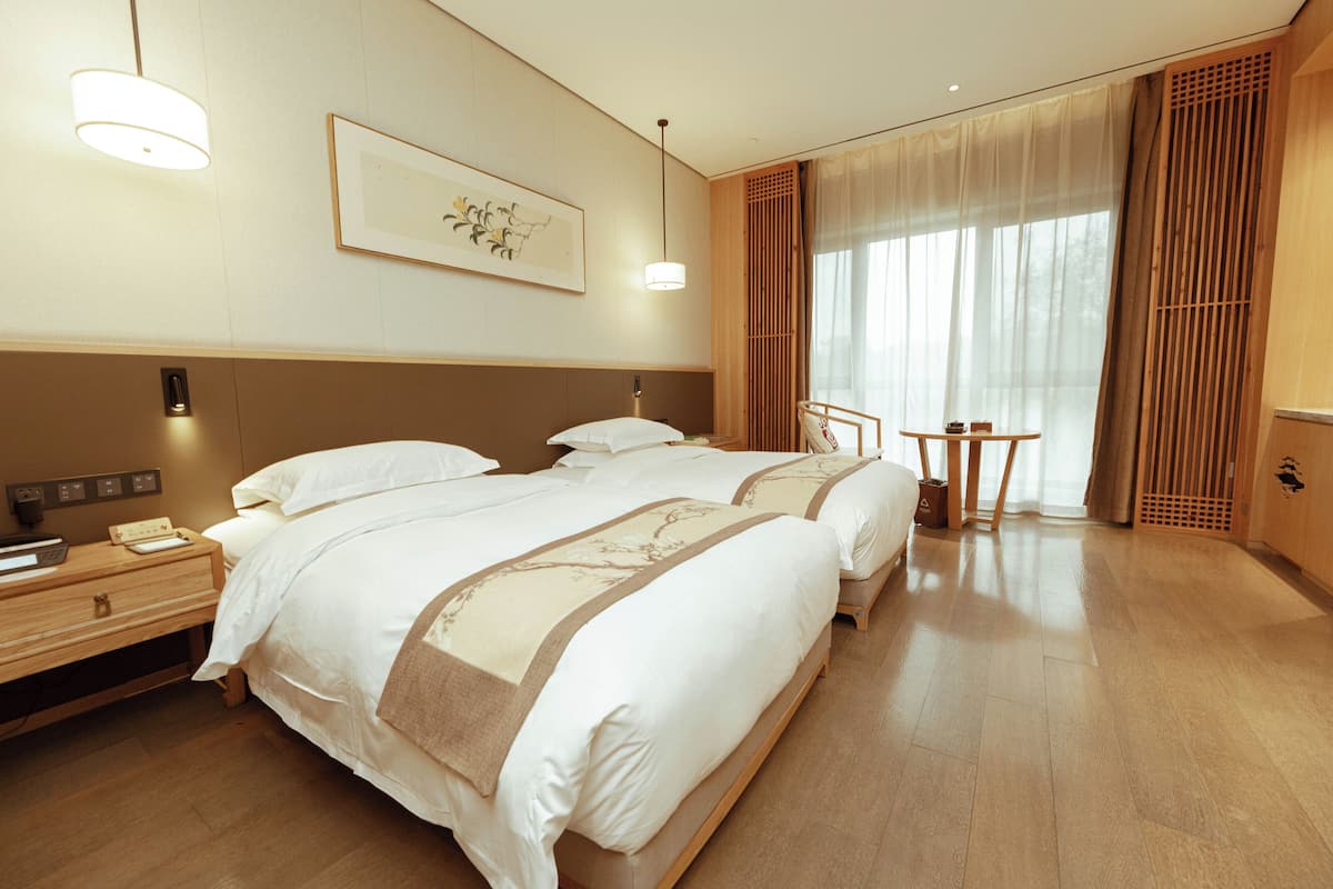 A hotel room with two beds, a bedside table, a lamp, a small table with a chair, and a large window with curtains.