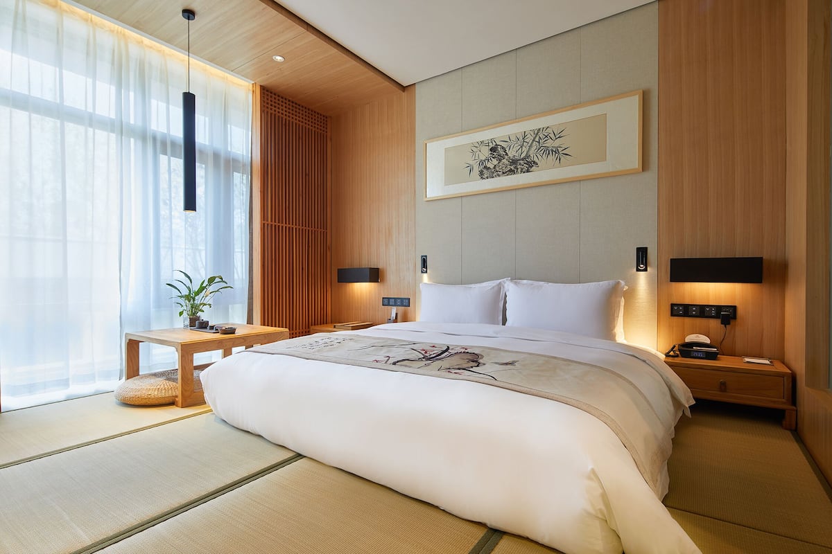 A hotel room with a large bed, bedside tables, a bedside lamp, a wall-mounted artwork, and a window with sheer curtains.