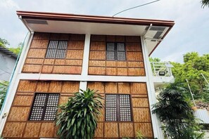 Un edificio de varios pisos con techo plano, ventanas grandes con celosías de madera y una unidad de aire acondicionado visible en el segundo piso.