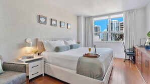 Ein modernes Hotelzimmer mit einem großen Bett, einem Nachttisch mit Lampe, einem Schreibtisch mit Stuhl und Blick auf die Stadt durch das Fenster.