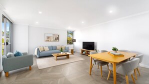 A modern living room with a sofa, coffee table, TV, and dining area. - Oceans Edge #25 | Top Floor,1 Min to Terrigal Beach| Accom Holidays (TERRIGAL)