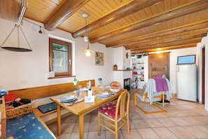 A dining area with a wooden table, chairs, and a bench. There is a kitchen in the background with a refrigerator and cabinets. - Rustico San Martin - Monti di Ditto (Cugnasco-Gerra)