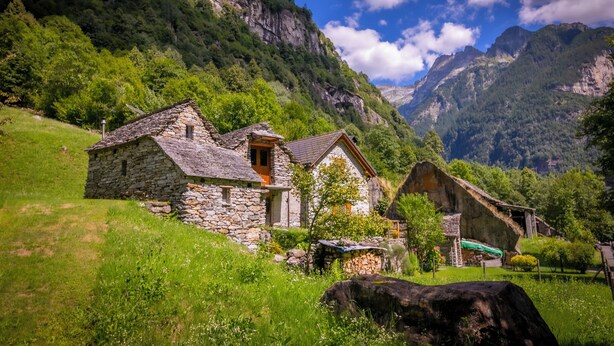 A stone cottage with a thatched roof, surrounded by lush greenery and mountains in the background. - Rustico San Martin With Garden in Monti di Dritto (Cugnasco-Gerra)