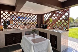 Un área de cocina exterior con mesa y sillas, una cafetera y vista al jardín.