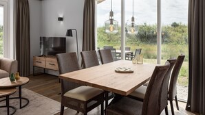A modern dining area with a wooden table and wicker chairs, a TV, and a view of greenery through large windows. - Villa in Someren Near Somerense Heide (Someren)