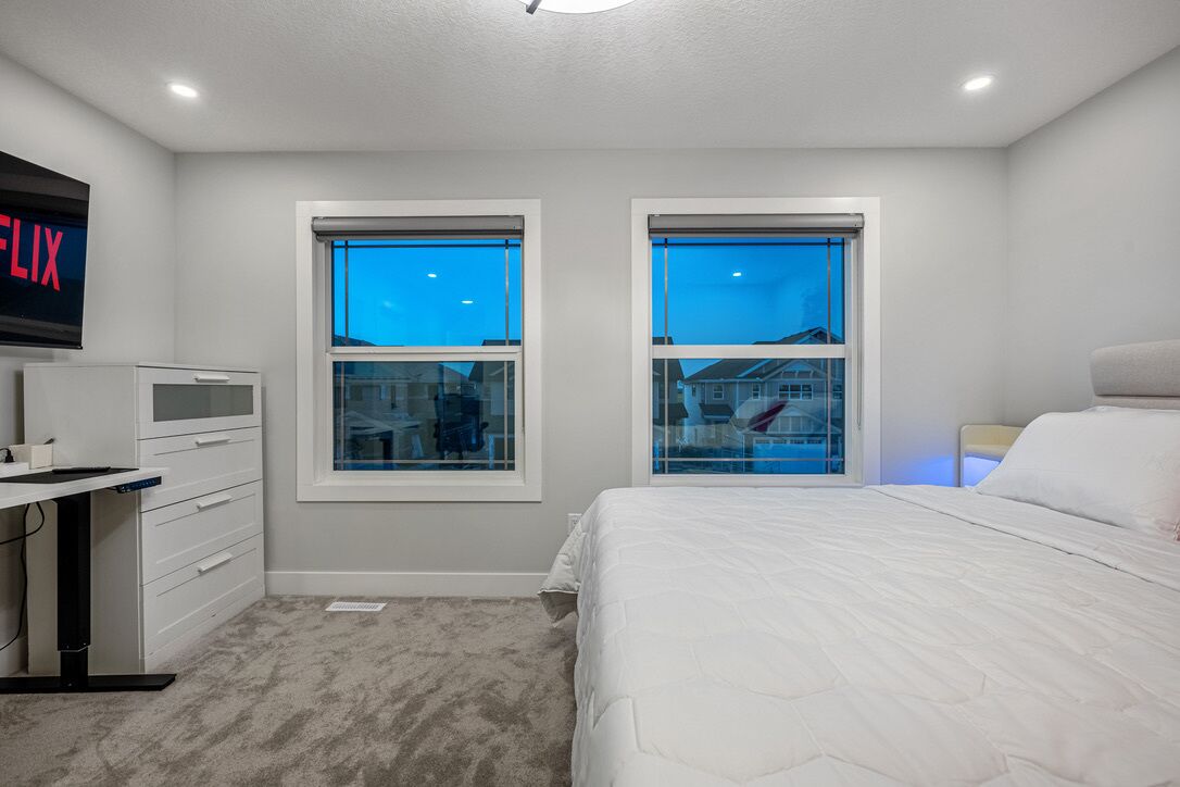 A bedroom with a bed, a desk, two windows, and a TV on the wall.