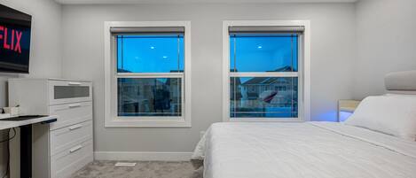 A bedroom with a bed, a desk, two windows, and a TV on the wall.