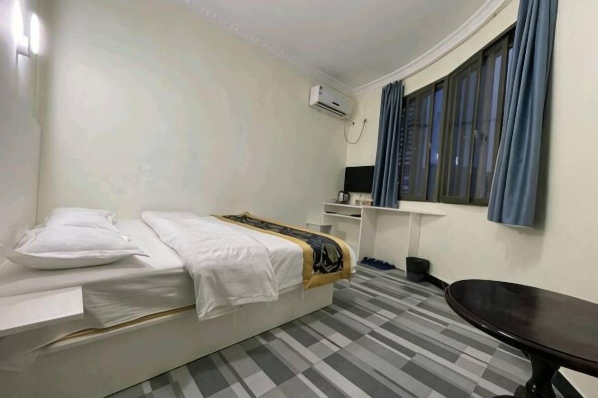 A hotel room with a bed, a desk, a chair, a window with curtains, and an air conditioning unit.
