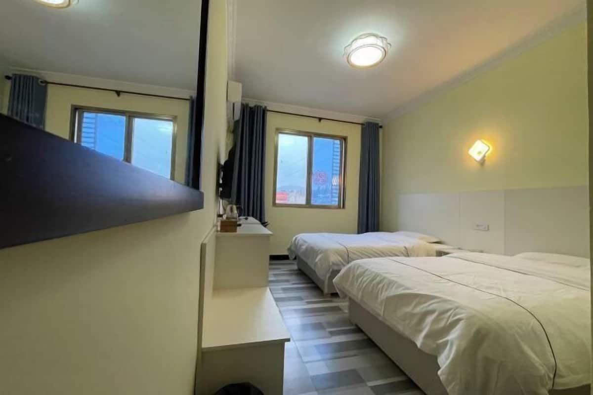 A hotel room with a bed, a desk, a chair, a window with curtains, and a wall-mounted light.