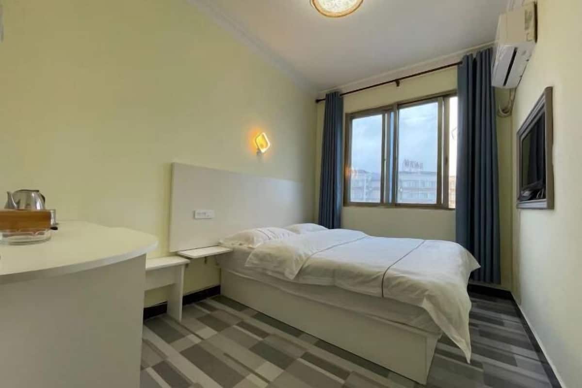 A hotel room with a bed, a bedside table, a small desk, and a window with curtains.