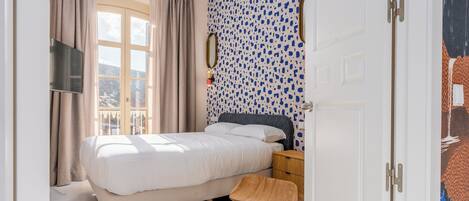 A bedroom with a bed, a wooden chair, a nightstand, a TV, and a window with curtains.