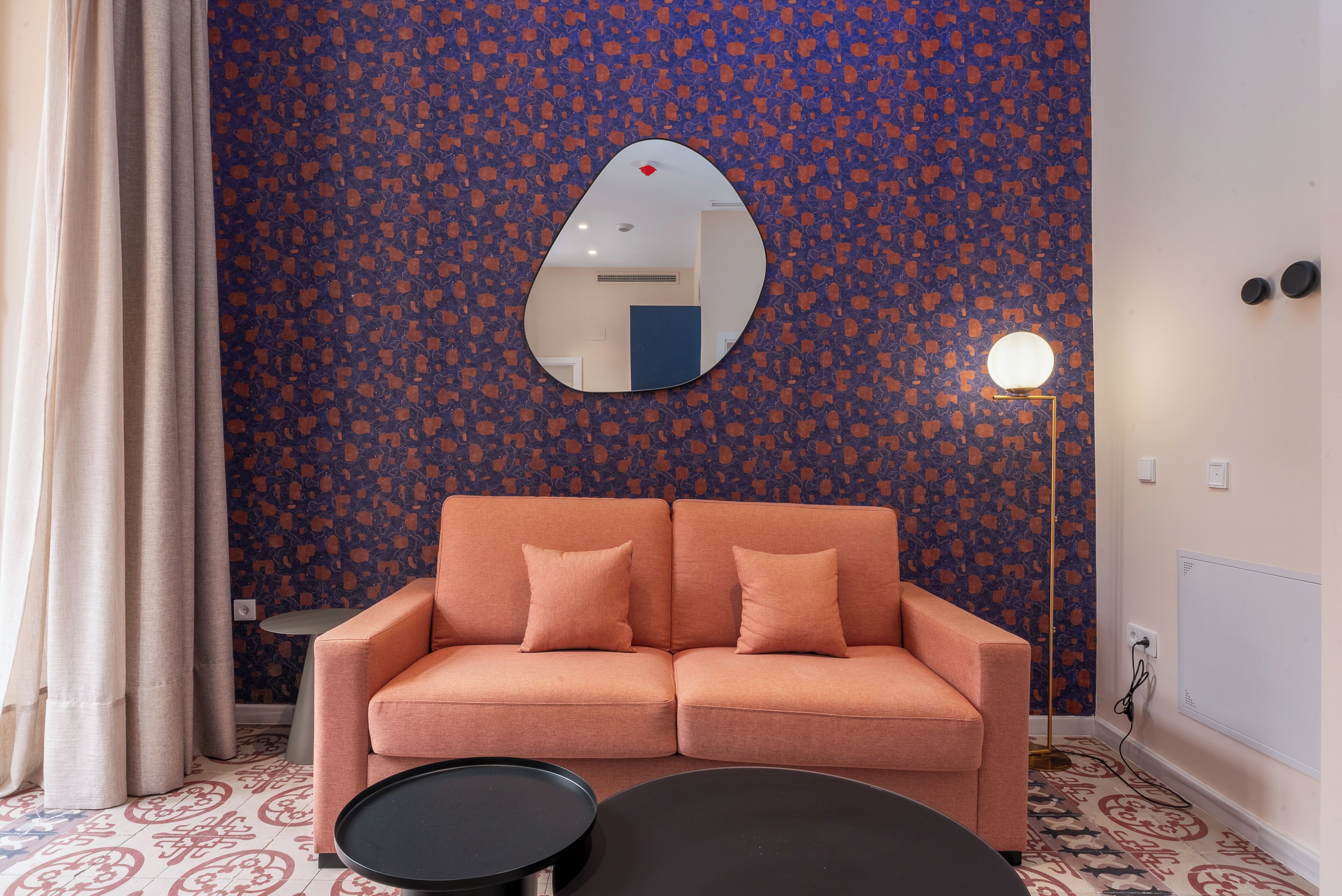 A room with a patterned wall, a sofa with cushions, a round table, a mirror, and a floor lamp.
