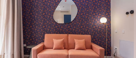A room with a patterned wall, a sofa with cushions, a round table, a mirror, and a floor lamp.